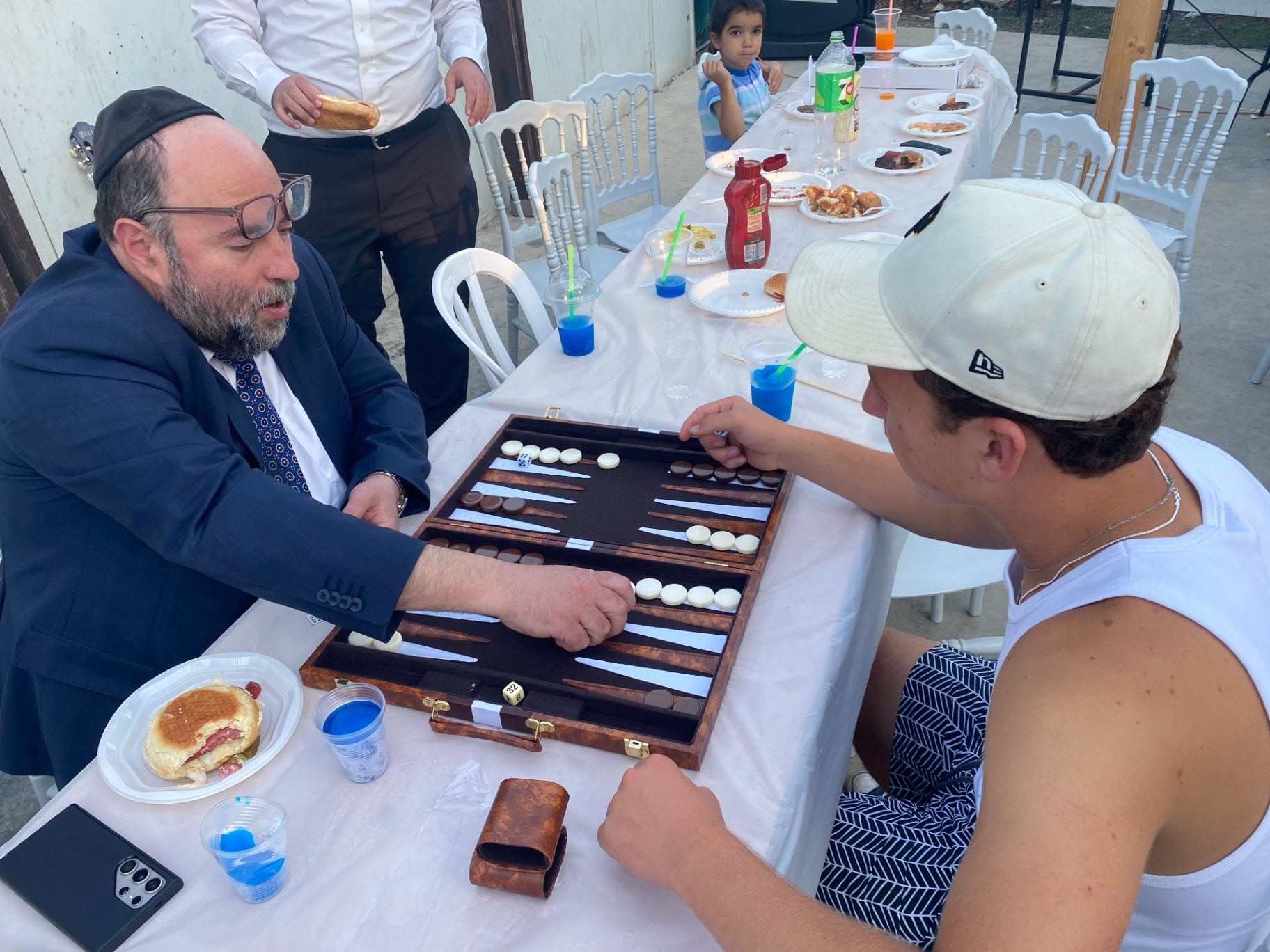 A rabbi and a talmid playing backgammon at a barbecue table
