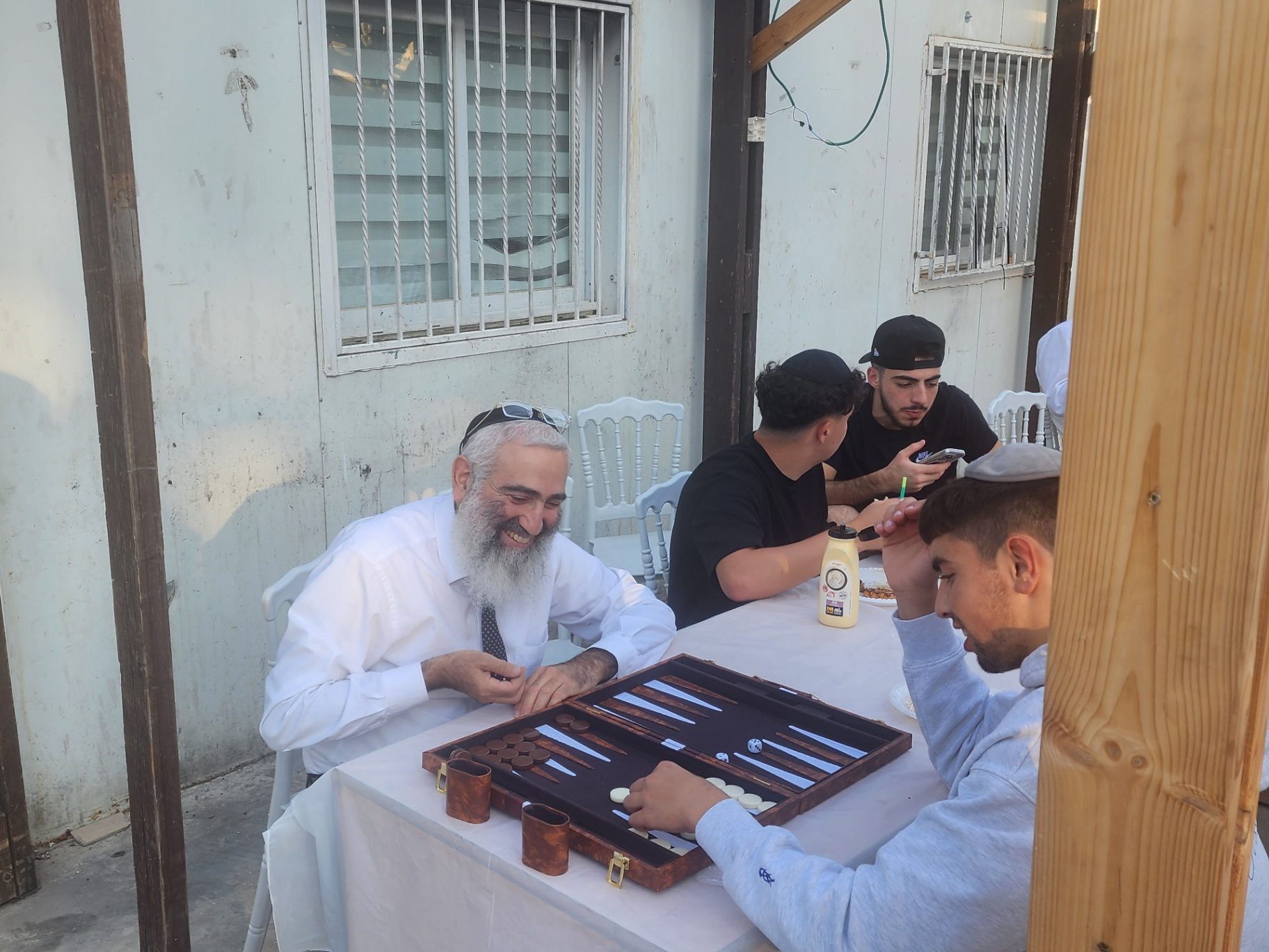 A rabbi laughing with talmidim over a backgammon board at an outdoor meal